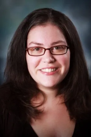 A woman with long dark hair and glasses smiles at the camera, wearing a black top against a blurred background.